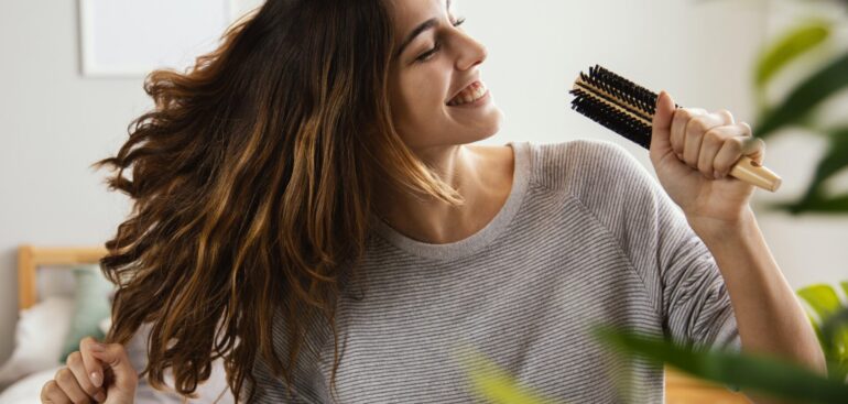 Mulher feliz, com cabelos longos e brilhantes, sorrindo e usando uma escova de cabelo como microfone. Ela está cantando e dançando em um quarto claro, com a mão livre levantada, expressando alegria e confiança.