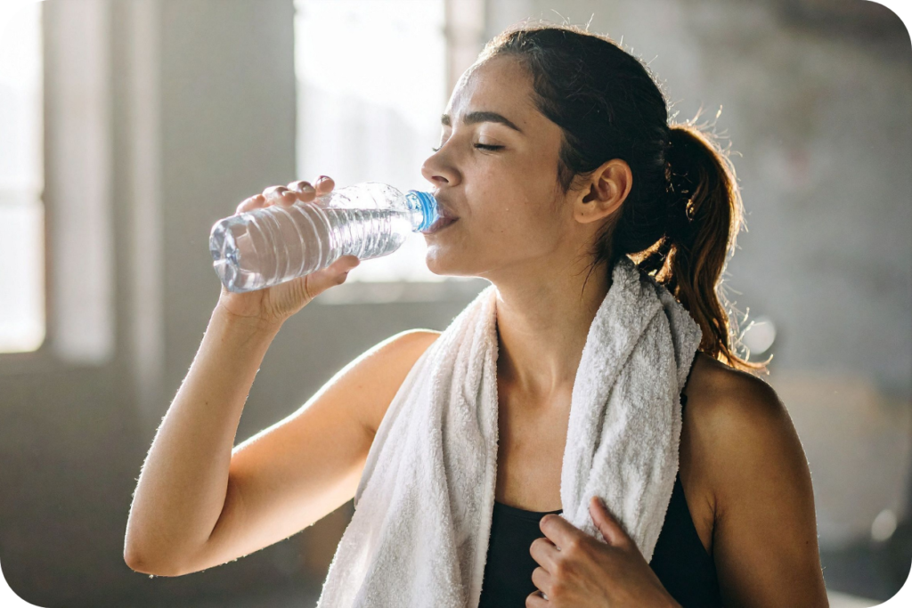 Mulher jovem, morena, com uma toalha branca no pescoço, bebendo água de uma garrafa plástica após um treino ou atividade física, em um ambiente interno com luz natural, demonstrando a importância da hidratação para a recuperação e bem-estar.