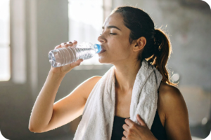 Mulher jovem, morena, com uma toalha branca no pescoço, bebendo água de uma garrafa plástica após um treino ou atividade física, em um ambiente interno com luz natural, demonstrando a importância da hidratação para a recuperação e bem-estar.